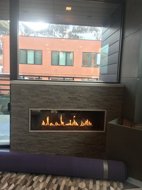 A linear fireplace in a stone wall beneath windows overlooking a snowy mountain