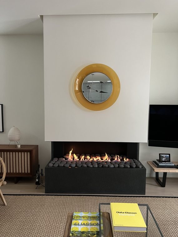 A contemporary fireplace with a pebble bed beneath a white surround and round yellow mirror